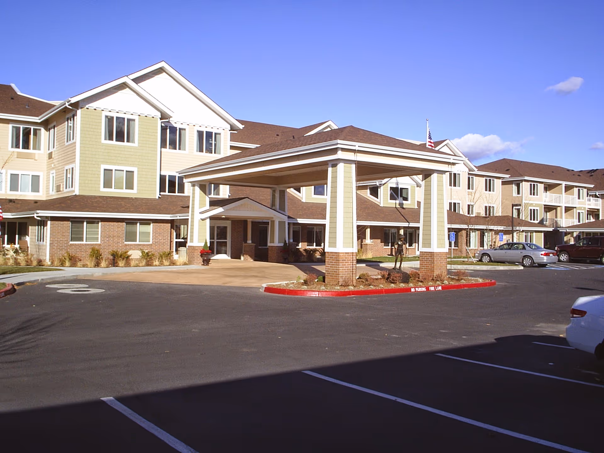 Exterior view of a multi-story senior living facility with a covered entrance, parking spaces, and a clear blue sky.