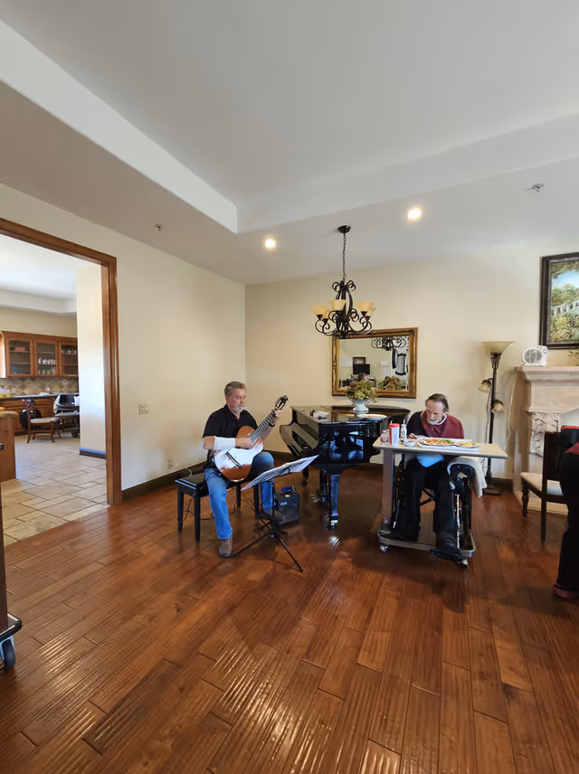 A man playing guitar seated on a bench next to a grand piano in a room with wooden floors and beige walls. Another man in a wheelchair with a tray table is nearby, eating a meal. The room has a chandelier, a mirror on the wall, a floor lamp, and a fireplace with a painting above it. An open doorway leads to another room with a dining table and chairs.