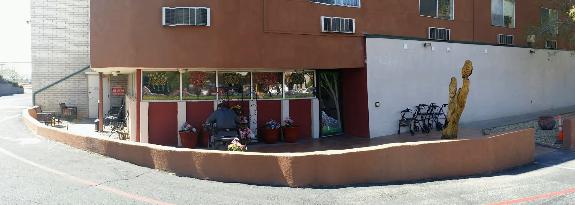 Exterior front entrance of a retirement apartment building with a covered patio, potted plants, seating, a person on a mobility scooter, several walkers, and a wooden sculpture.