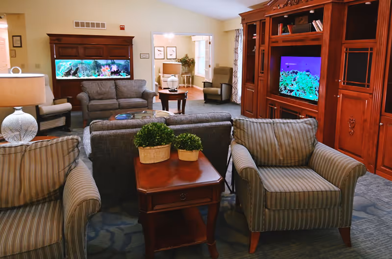 A cozy living room area in a senior living facility featuring two striped armchairs, a gray sofa, a wooden side table with two potted plants, a large wooden entertainment center with a TV displaying an aquarium scene, and a built-in fish tank. In the background, there is another seating area with armchairs and a lamp.