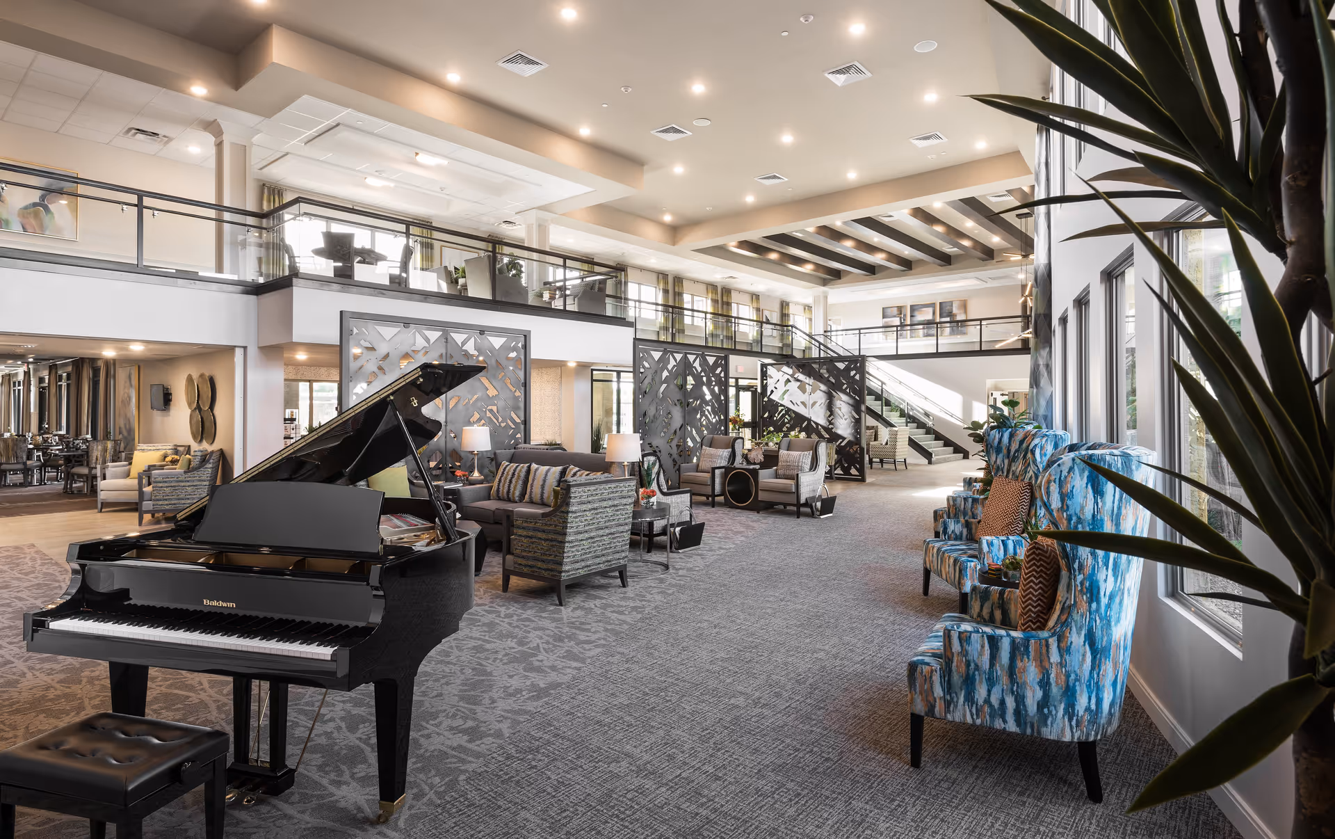Spacious and well-lit senior living facility common area with a black grand piano in the foreground, comfortable seating including patterned armchairs and sofas, decorative room dividers, large windows allowing natural light, and a mezzanine level with additional seating and a staircase.