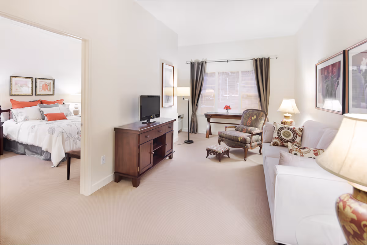 A bright and cozy living room with a white sofa adorned with patterned pillows, a floral upholstered armchair with a matching footstool, a wooden TV stand with a flat-screen TV, a floor lamp, and a window with dark curtains. Adjacent to the living room is a bedroom with a bed dressed in white and red bedding, two framed pictures above the bed, and a bedside lamp.