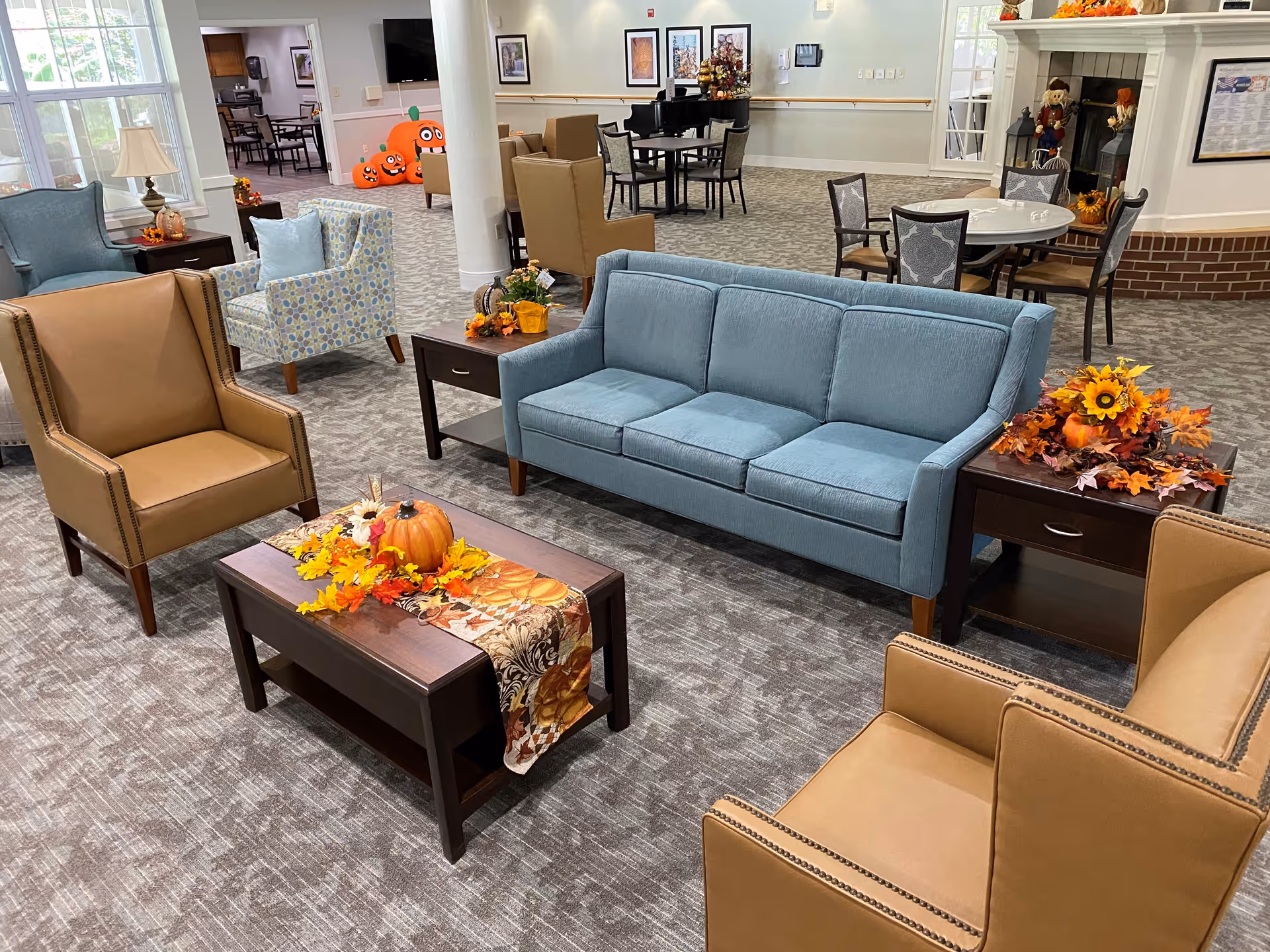 A cozy senior living facility common area with a blue sofa, two tan armchairs, and a patterned armchair arranged around a wooden coffee table decorated with autumn-themed items including a pumpkin and fall leaves. In the background, there are additional seating areas with tables and chairs, a fireplace with seasonal decorations, and a piano. The room has carpeted floors and large windows letting in natural light.