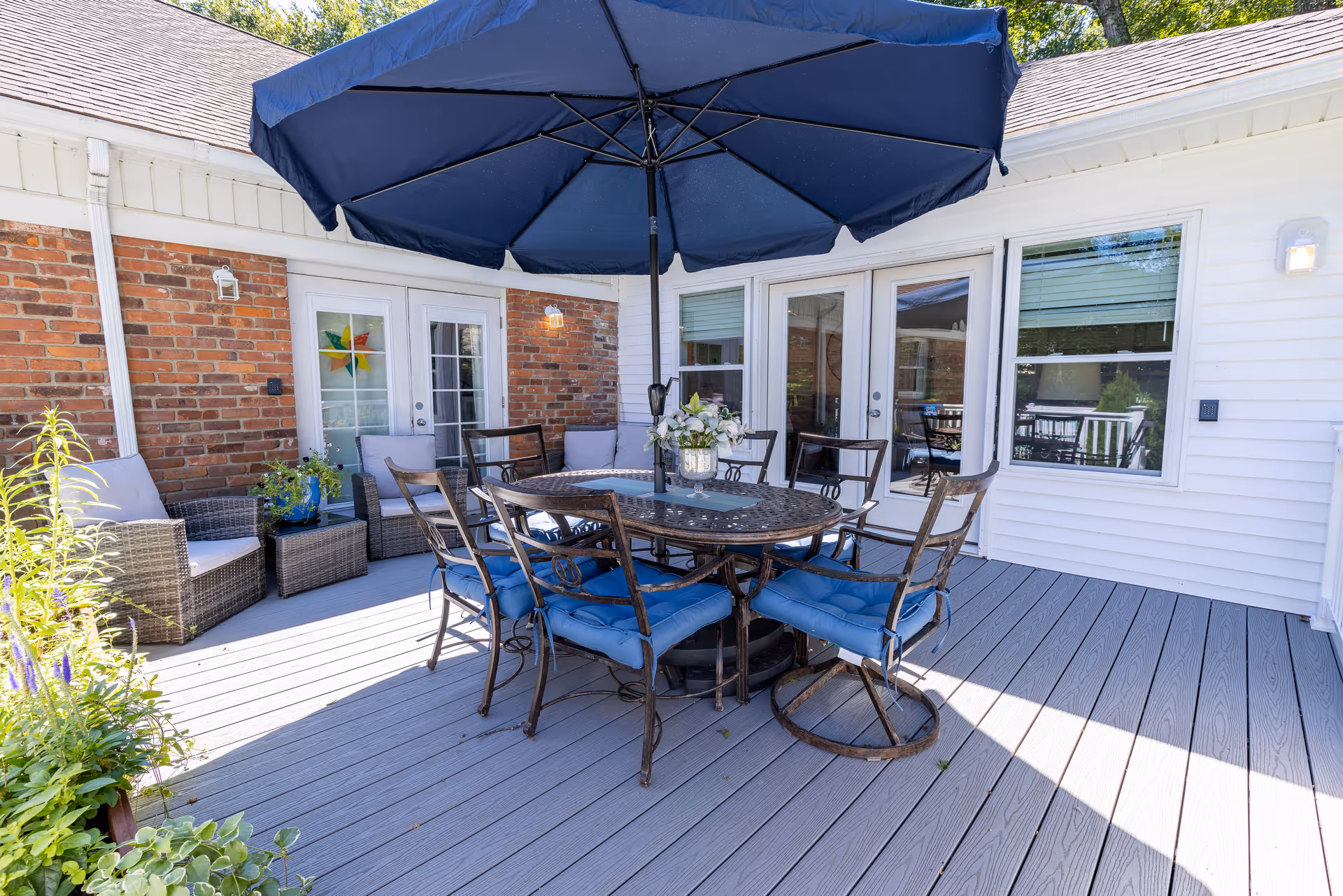 Outdoor deck patio with a round dining table, blue umbrella and cushioned chairs in front of a brick-and-siding building.