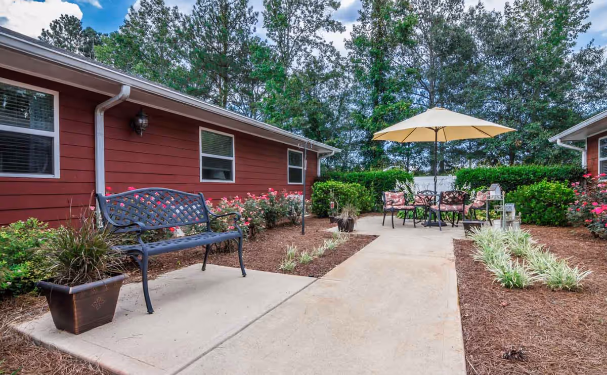 Outdoor patio area at Oaks at Post Road featuring a black metal bench, a potted plant, a concrete walkway, and a round table with four chairs under a large beige umbrella surrounded by greenery and red buildings.
