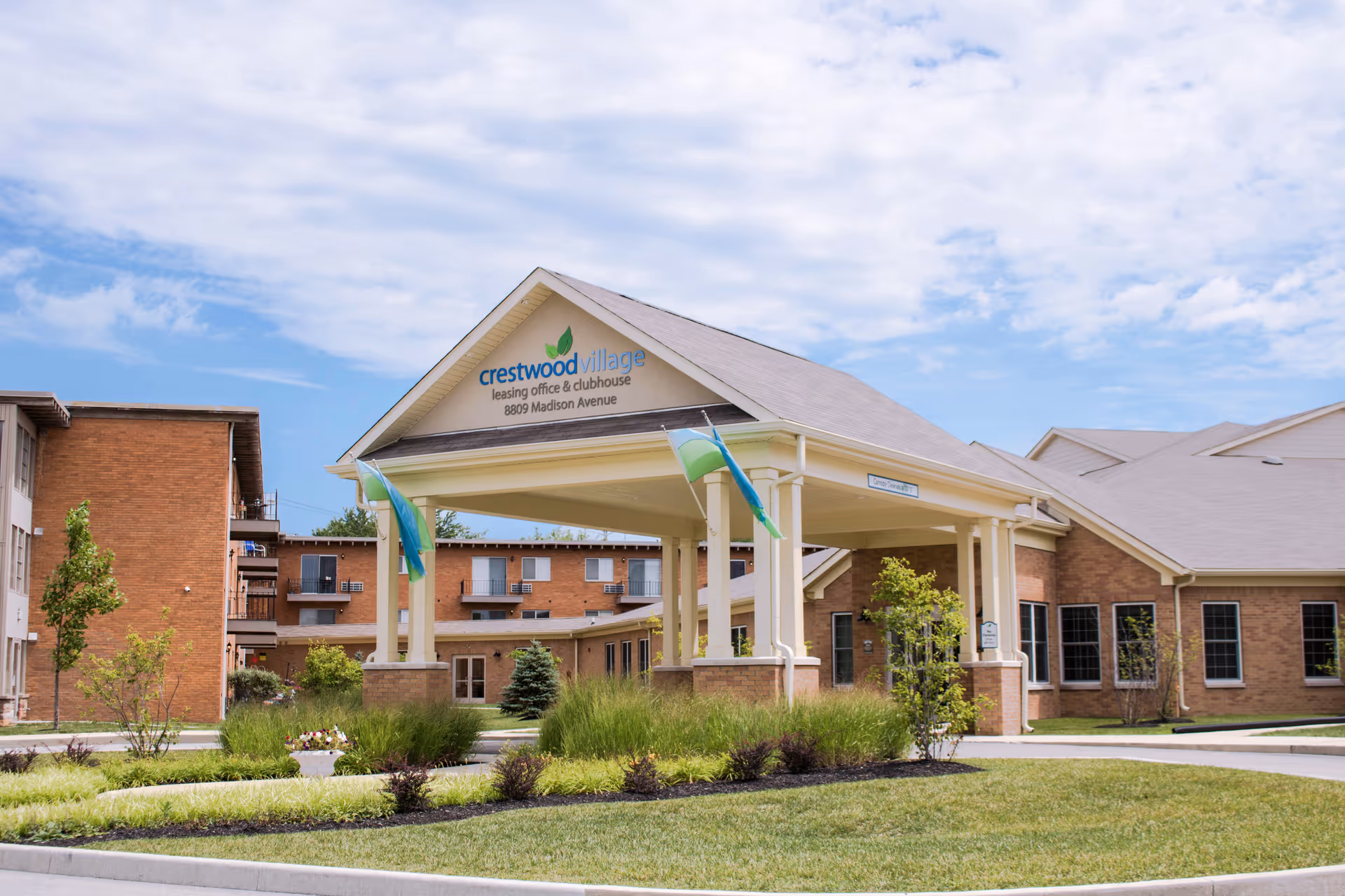Front entrance of Crestwood Village Senior Community showing a covered porte-cochere, flags, and surrounding landscaping in front of a brick building.