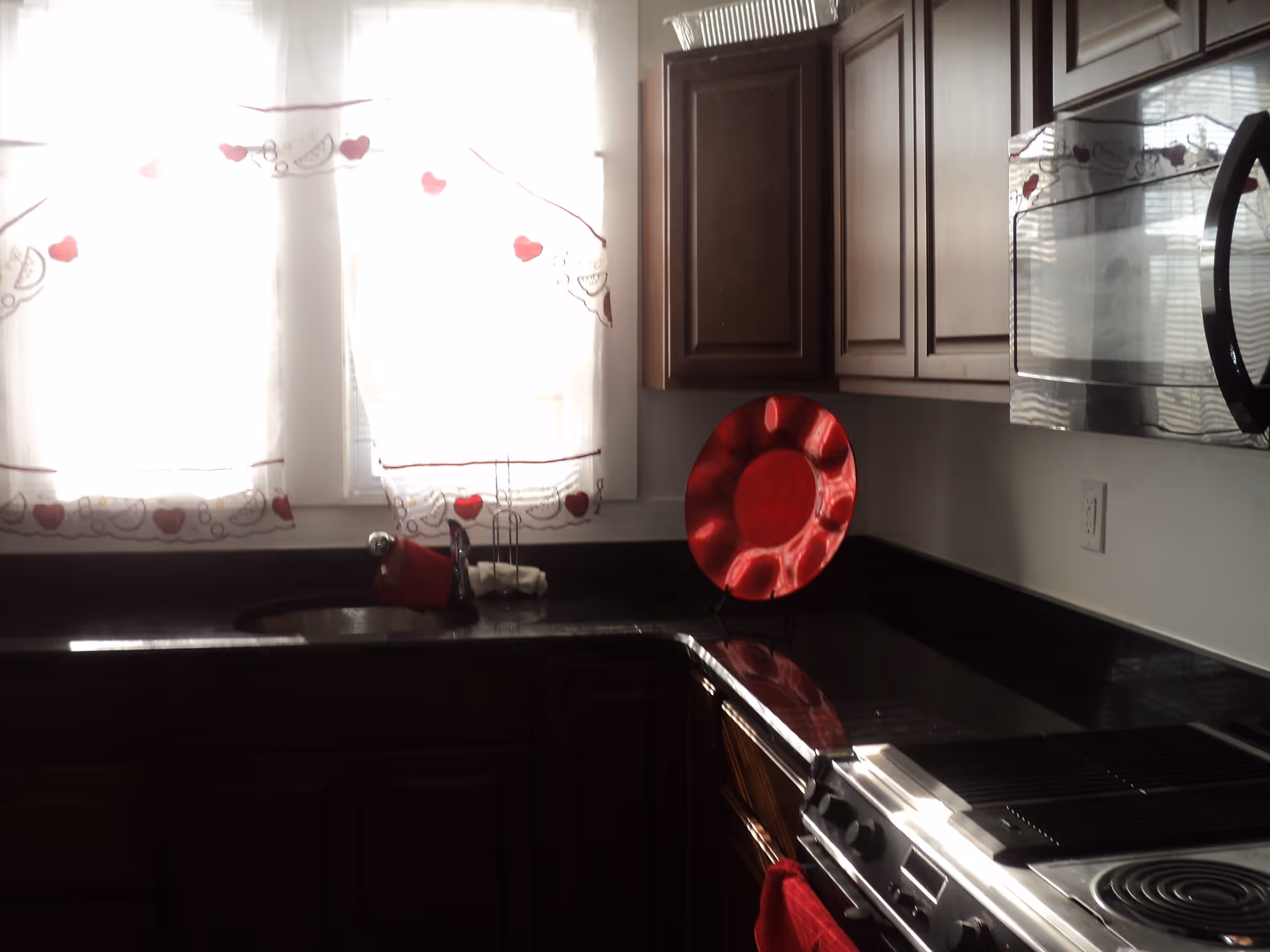 A kitchen with dark wooden cabinets, a black countertop, a stainless steel stove and microwave, a sink under two windows with white curtains decorated with red hearts, and a large decorative red plate on the counter.