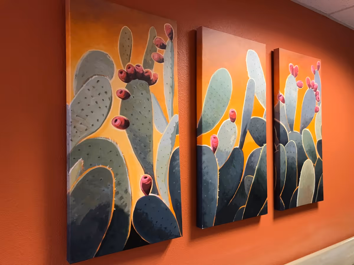 Three colorful cactus canvas paintings hung on an orange wall in a hallway.