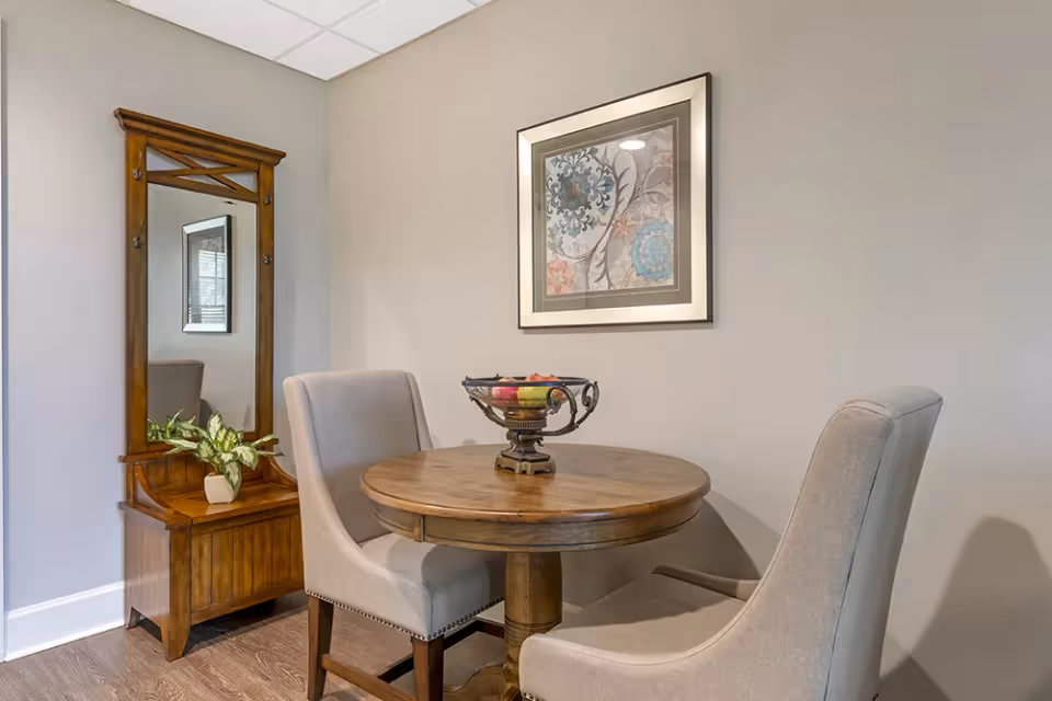Small dining nook with a round wooden table, two upholstered chairs, a decorative fruit bowl, framed artwork on the wall, and a wooden hall tree with a mirror.