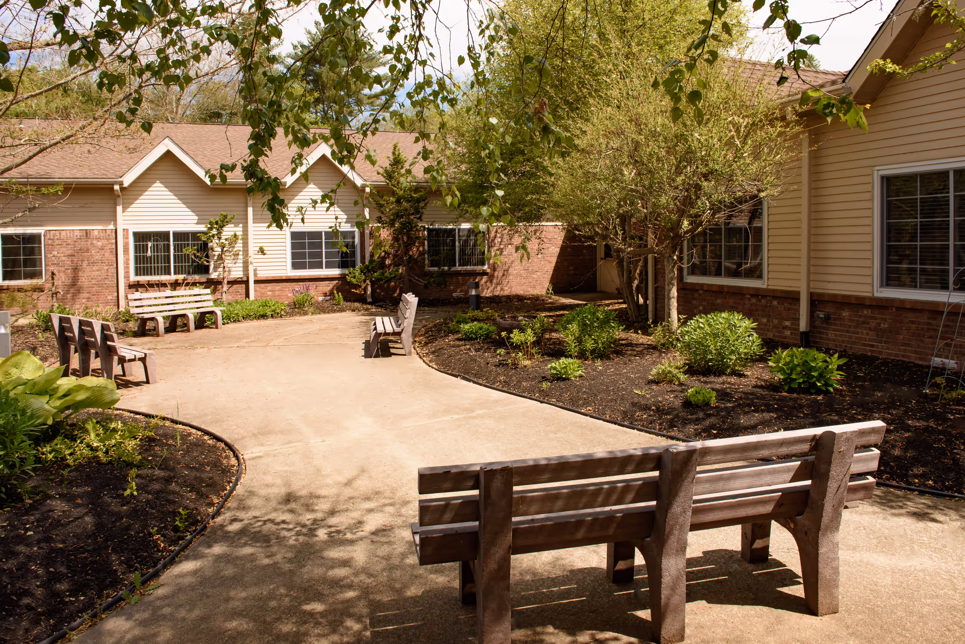Outdoor courtyard area at The Oaks facility with several wooden benches arranged along a curved concrete pathway. The courtyard is surrounded by garden beds with small shrubs and trees, and the building exterior features brick and beige siding with multiple windows.