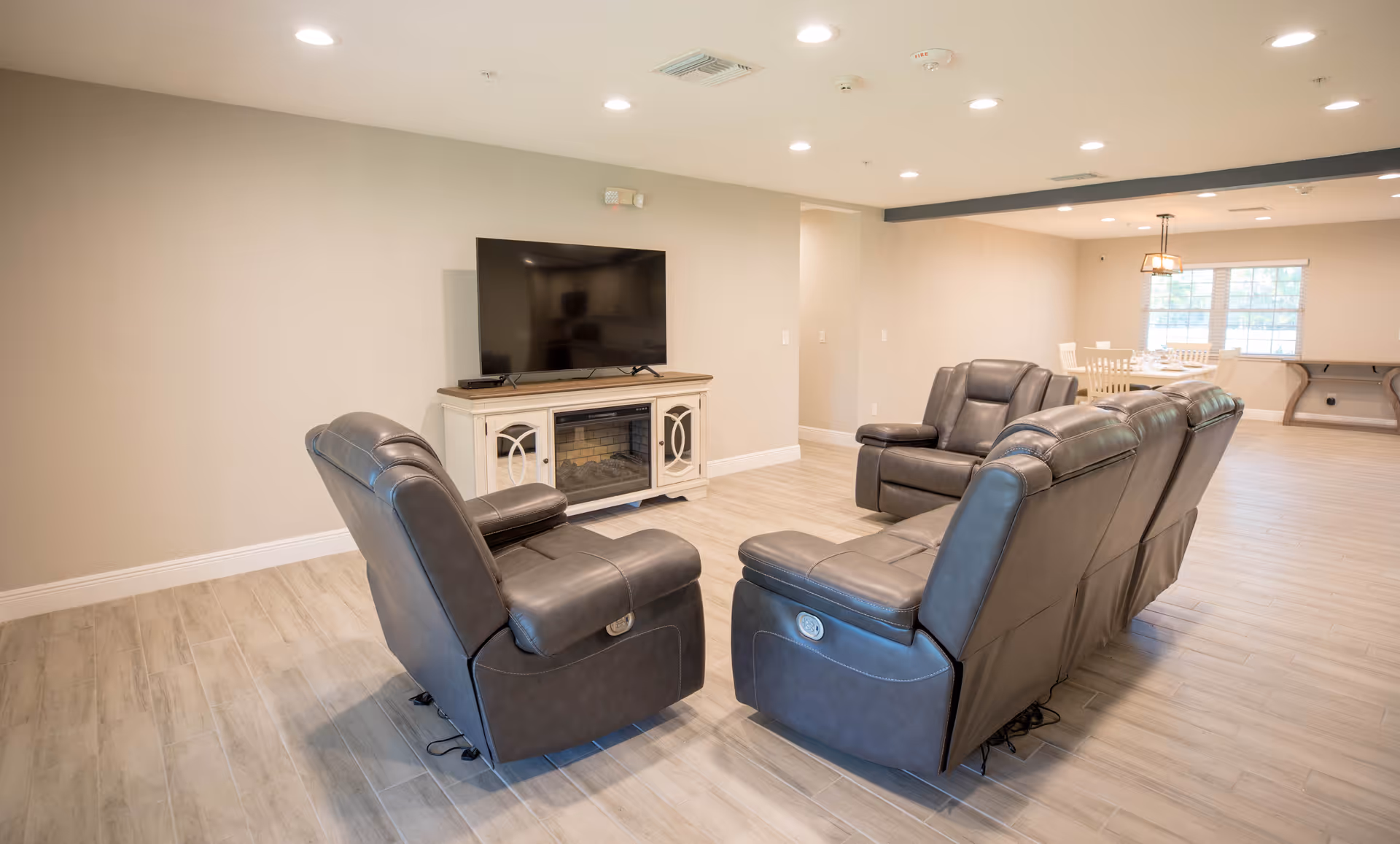 A spacious living room area with four dark leather recliner chairs arranged around a TV mounted on a white cabinet with a built-in electric fireplace. The room has light wood flooring, beige walls, recessed ceiling lights, and a dining area with a table and chairs near a window in the background.