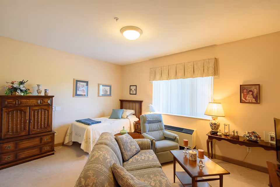 A cozy senior living bedroom with a single bed covered with a white quilt, a wooden headboard, and a blue throw blanket. The room includes a floral-patterned sofa, a comfortable armchair, a wooden coffee table with candles, a wooden cabinet, and a side table with a decorative lamp. The walls are adorned with framed pictures, and a large window with vertical blinds allows natural light to fill the room.