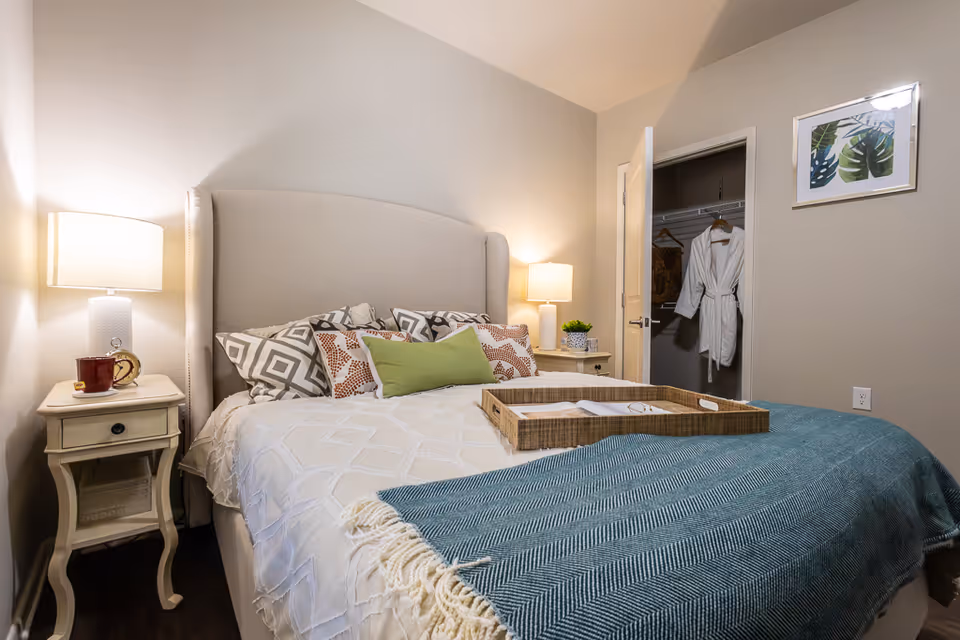 A cozy bedroom with a neatly made bed featuring multiple decorative pillows and a green throw blanket. There are two bedside tables with lamps on each side of the bed. One bedside table has a red cup and an alarm clock. The closet door is open, showing a white robe and some hanging clothes inside. A framed botanical print hangs on the wall next to the closet.