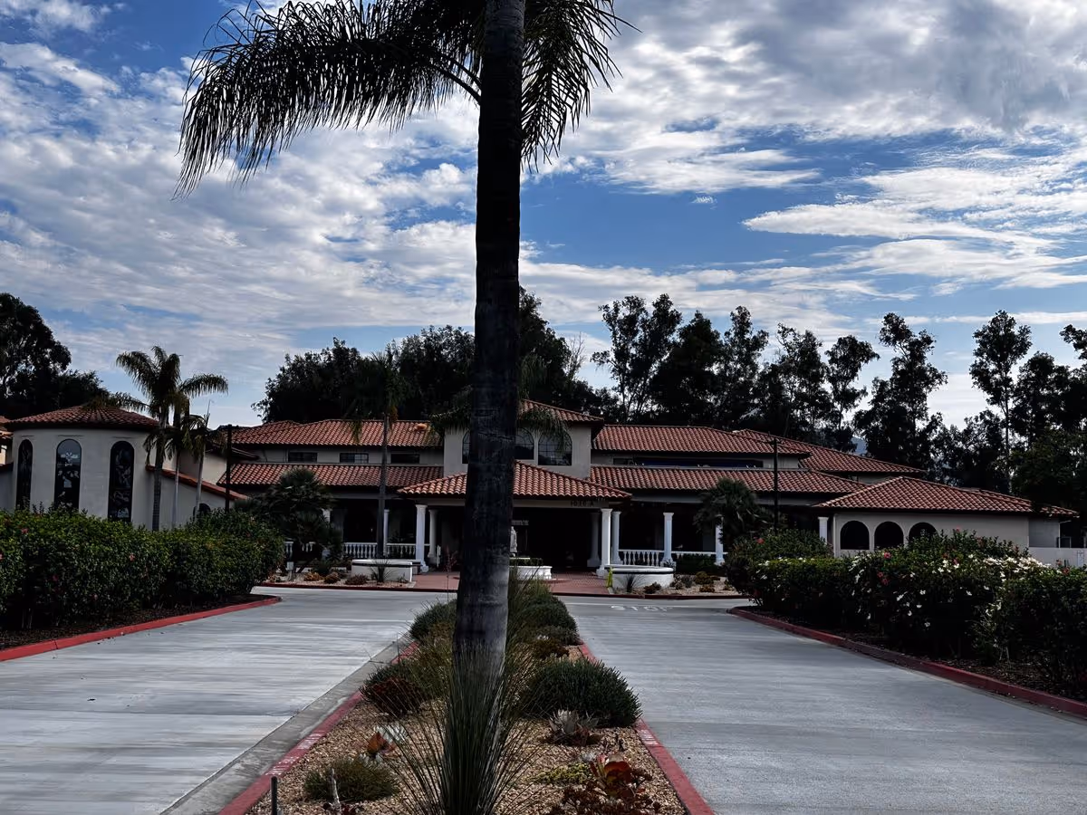Front exterior of a Mediterranean-style building with a red tile roof, palm trees, and a central driveway under a partly cloudy sky.