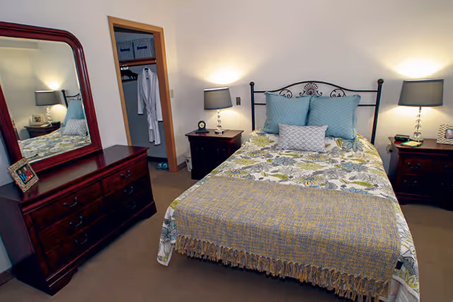 A neatly made bed with patterned bedding and throw in a furnished bedroom with nightstands, lamps, a dresser with mirror, and an open closet.