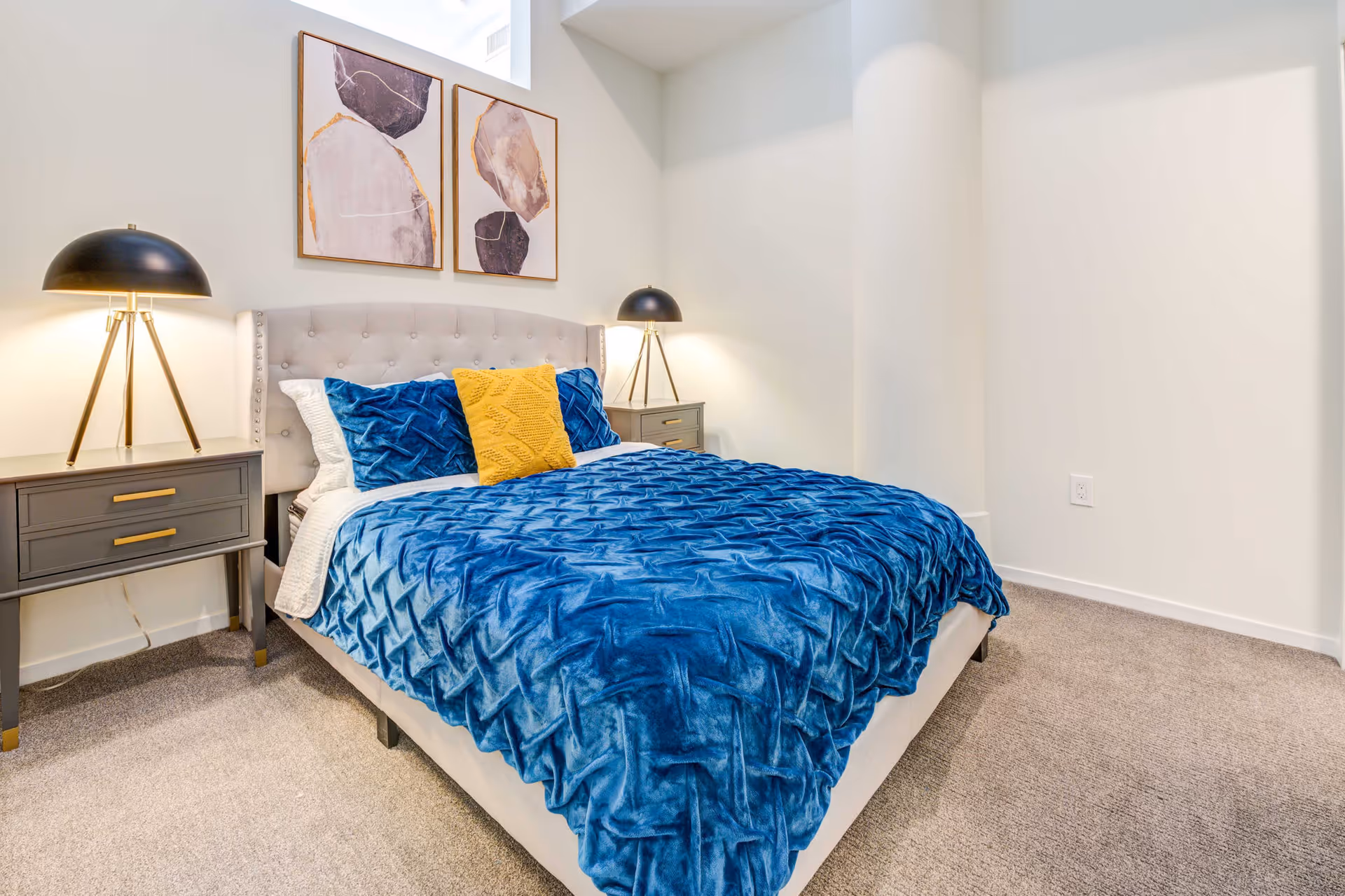A neatly made bed with a tufted light gray headboard, covered with a textured blue blanket and two blue pillows with a yellow pillow in the center. On each side of the bed are gray nightstands with gold handles, each topped with a black and gold tripod lamp. Two abstract art pieces hang on the white wall above the bed. The room has beige carpeting and white walls.