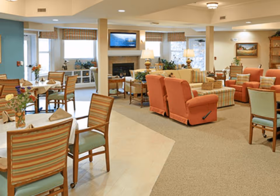 Open senior living common area with dining tables and striped chairs, and a seating area of upholstered sofas and recliners around a fireplace and TV.