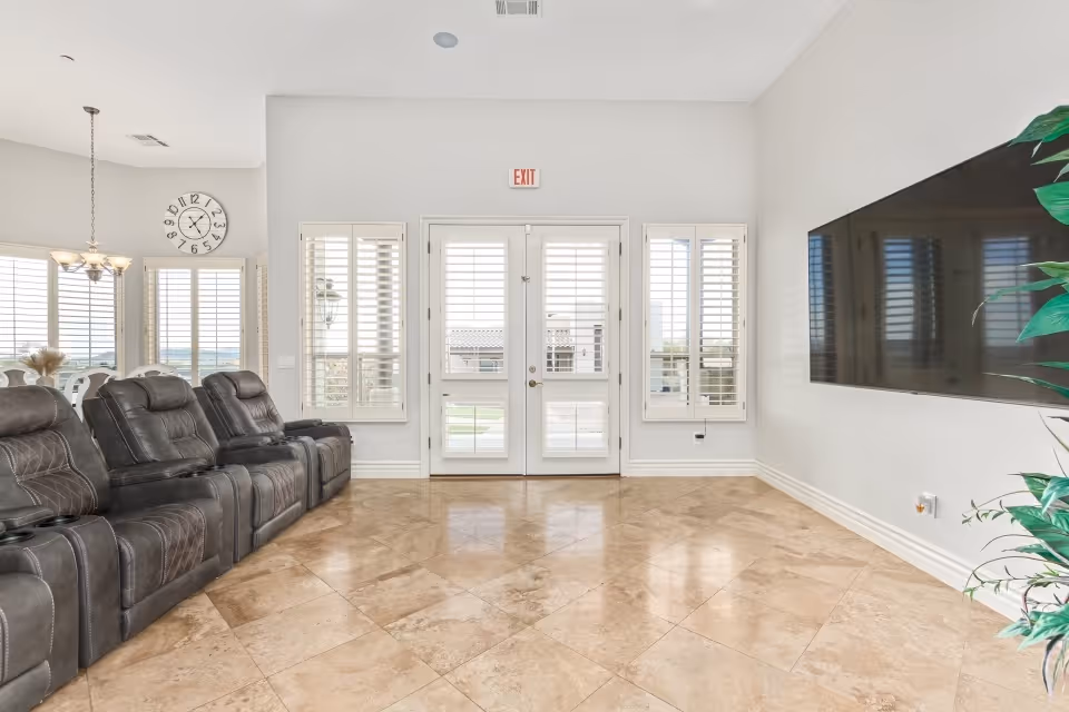A bright and spacious living room with beige tiled floors, a row of dark brown leather recliner chairs on the left, large windows with white shutters, double glass doors with an exit sign above, a large flat-screen TV mounted on the right wall, and a green potted plant in the corner.