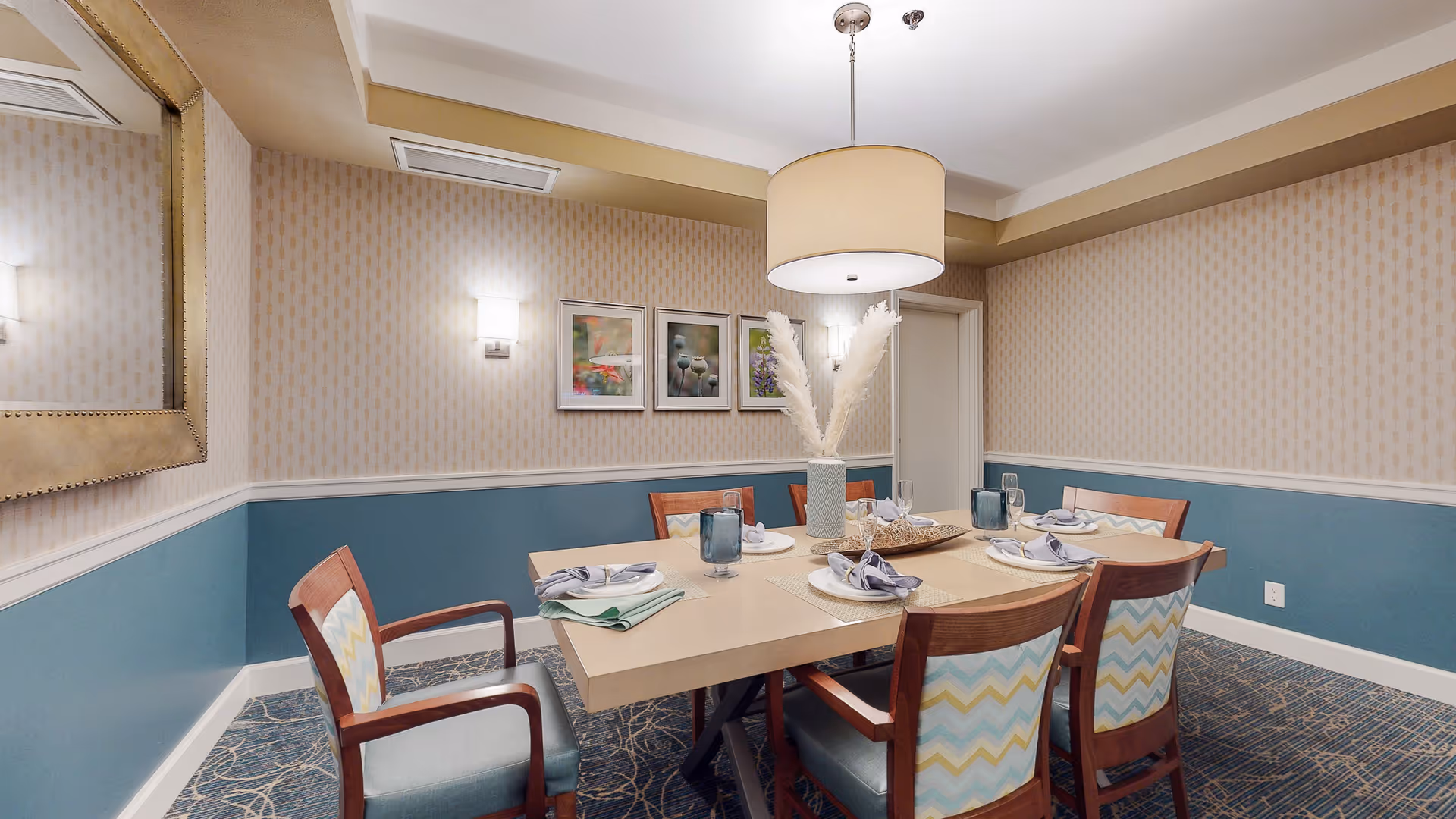 A dining room with a rectangular table set for six people. The table has placemats, folded napkins, plates, glasses, and a decorative vase with white pampas grass in the center. The chairs have wooden frames with patterned upholstery in blue, yellow, and white. The walls are painted blue on the lower half and beige with a subtle pattern on the upper half, adorned with three framed pictures and a large mirror. A round ceiling light fixture hangs above the table.