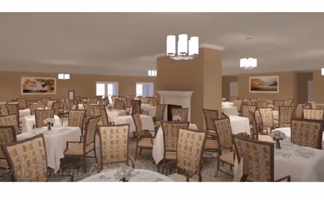 A spacious dining room with multiple tables covered in white tablecloths, each surrounded by patterned chairs. The room features beige walls, several large windows, framed artwork, and modern ceiling light fixtures. There is a fireplace in the center of the room.