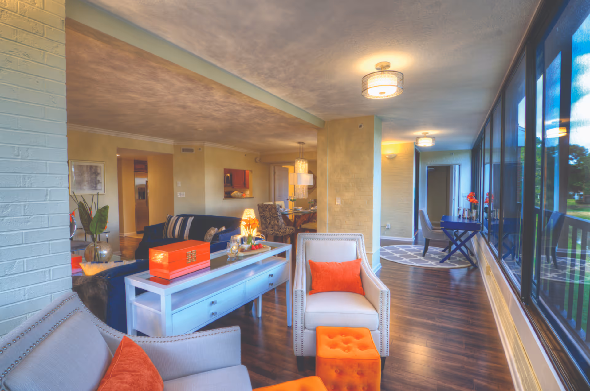 Bright open living area with upholstered chairs and orange accents, a console table, dining area in the background, and large floor-to-ceiling windows along one side.