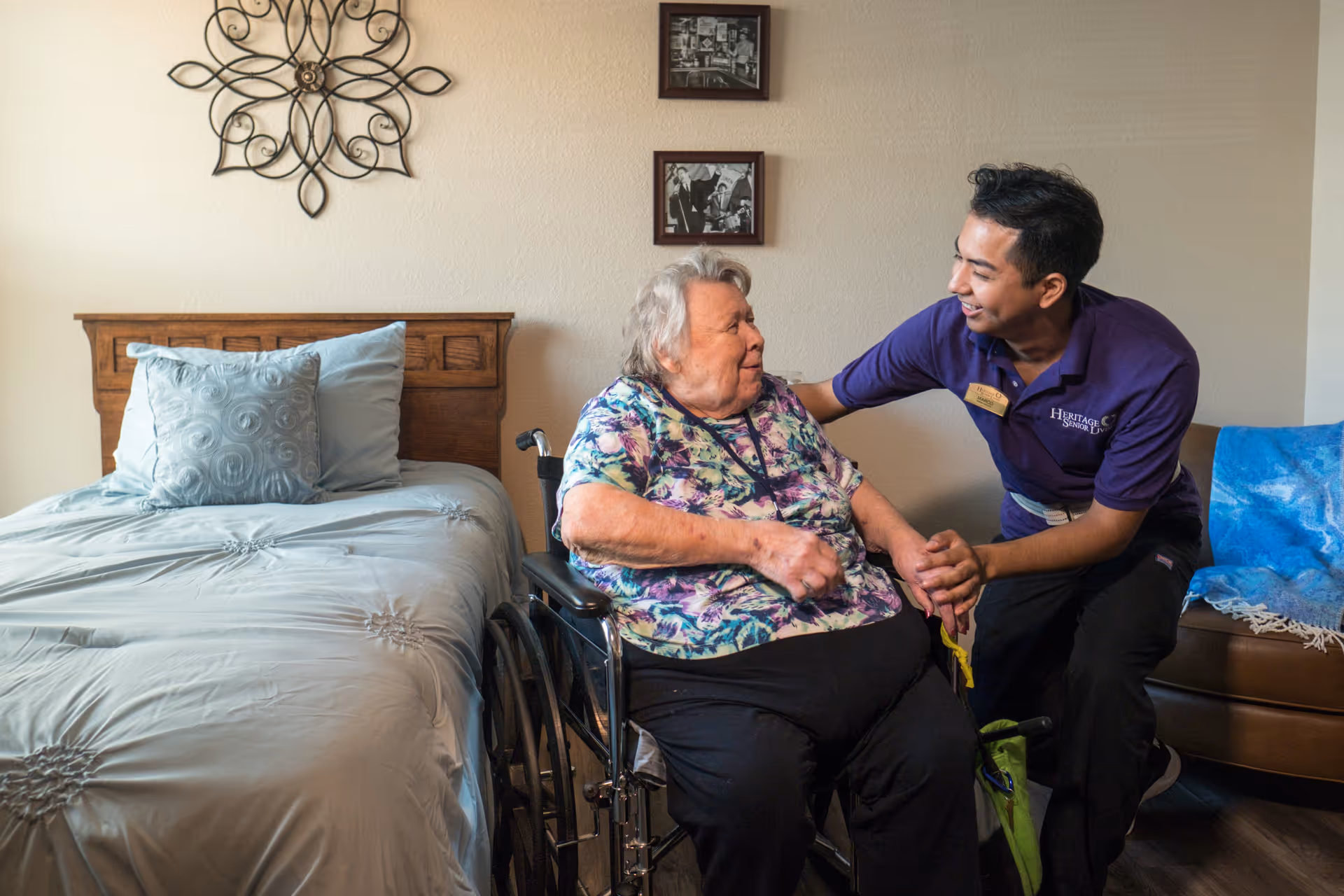 An elderly woman in a wheelchair is smiling and holding hands with a caregiver wearing a purple Heritage Senior Campus shirt in a bedroom with a bed, pillows, and wall decoration.