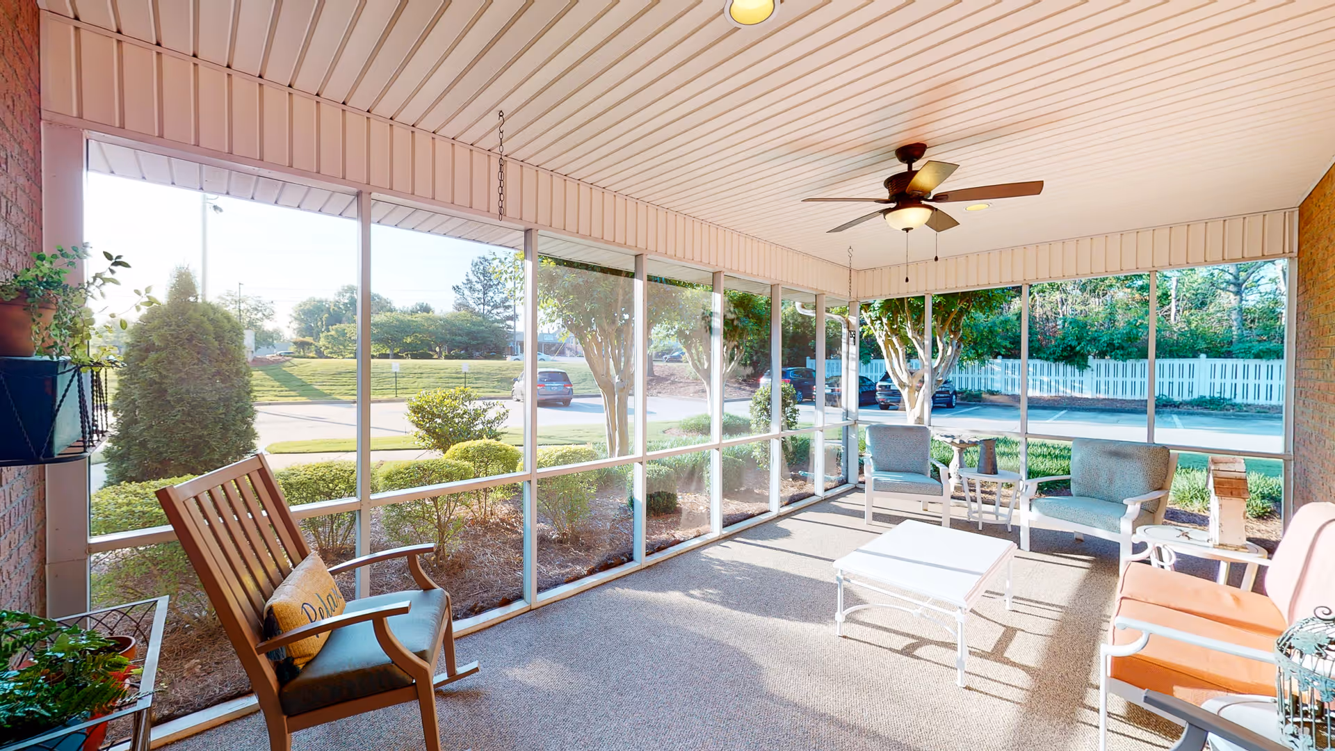 A bright and airy screened-in porch area with comfortable seating including cushioned chairs and a rocking chair. The porch has a ceiling fan with a light, potted plants, and a white coffee table. Outside the porch, there is a view of a parking lot, trees, and a white picket fence.