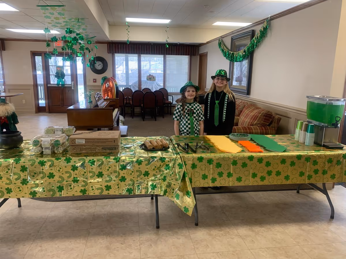 Two children stand behind a long table covered in green shamrock decorations and snacks in a community room decorated for St. Patrick's Day.