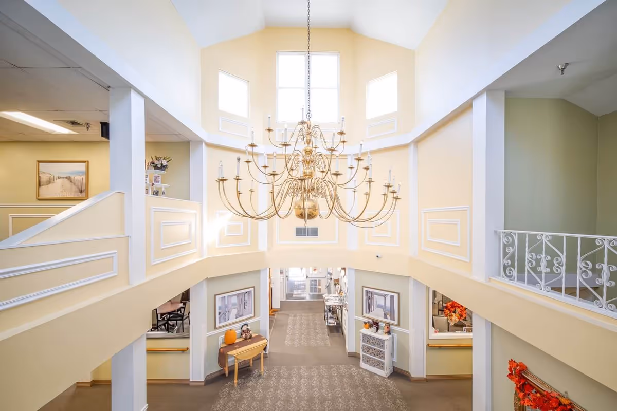 Interior view of a senior living facility lobby with high ceilings, large windows, and a grand chandelier hanging in the center. The walls are painted in soft yellow and cream tones with decorative molding. There are framed pictures on the walls, a small table with seasonal decorations, and a railing on the upper level overlooking the lobby.