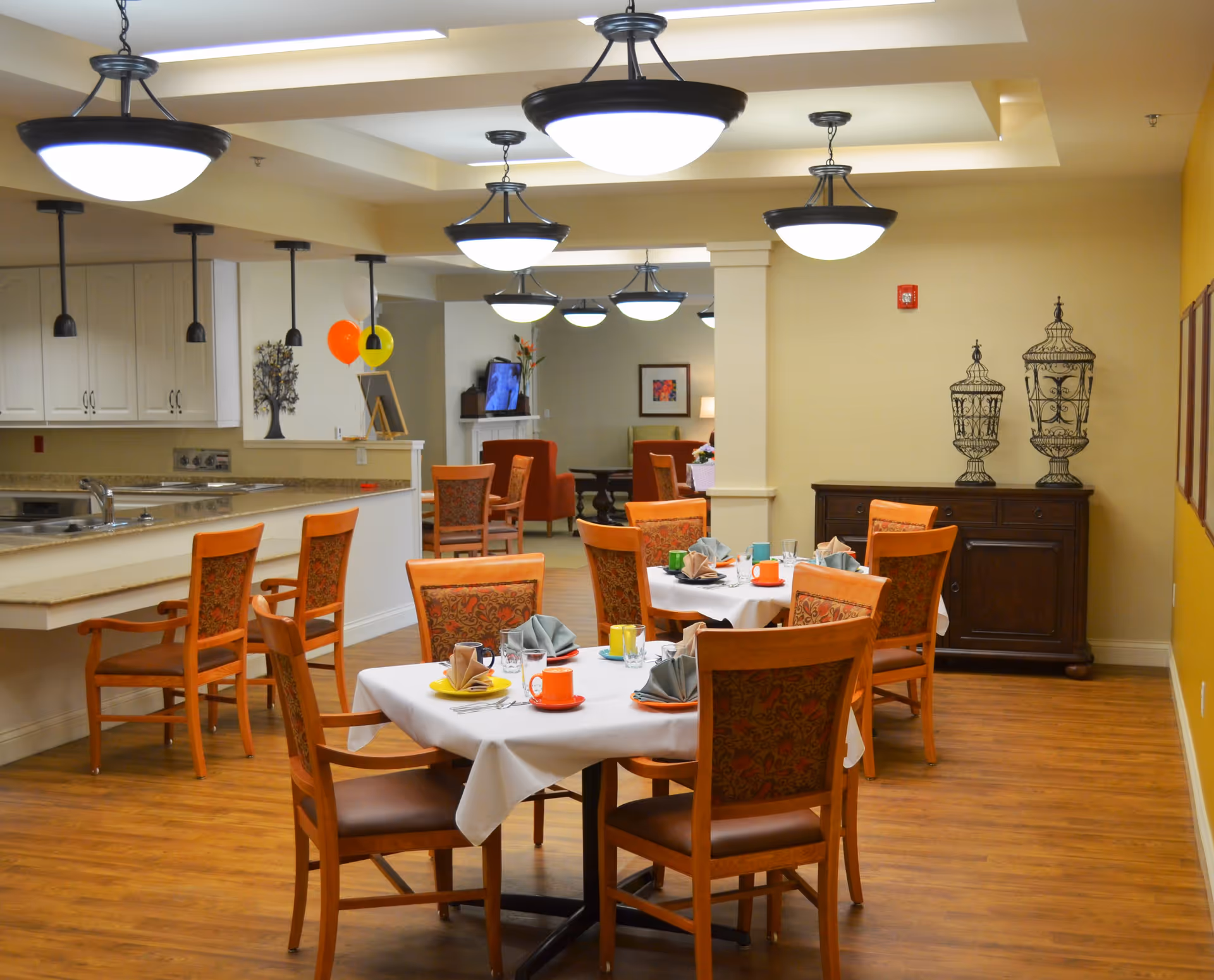 A dining area in Pinnacle Place Memory Care featuring wooden tables with white tablecloths, set with colorful cups and folded napkins. The room has wooden chairs with patterned upholstery, pendant lights hanging from the ceiling, and a kitchen counter with bar stools on the left. In the background, there is a living area with armchairs and a TV, and a wooden cabinet with decorative wire sculptures on the right.