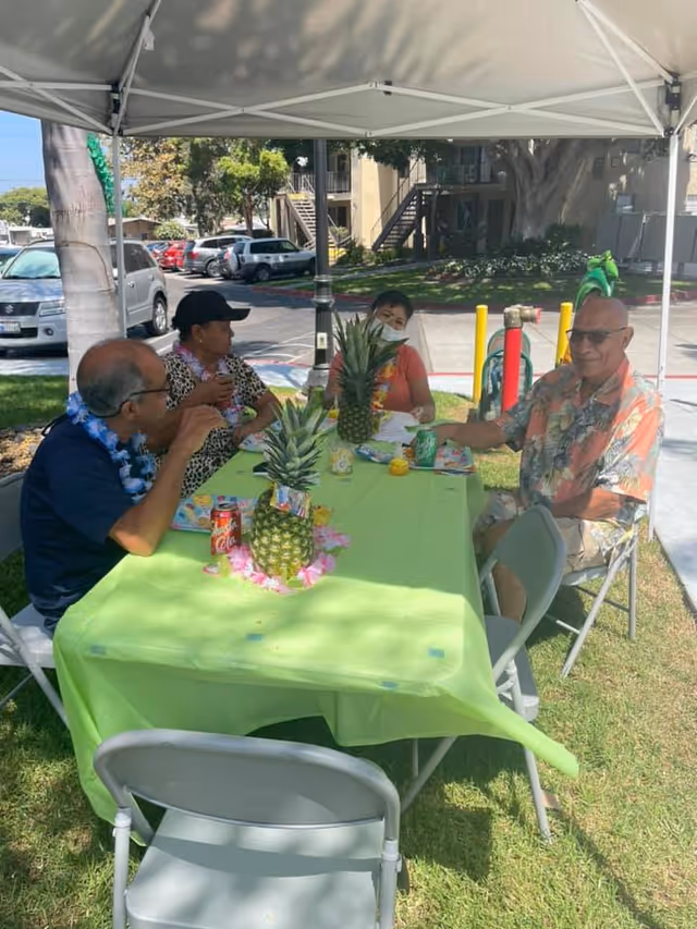 Four elderly people sitting around a green tablecloth-covered table under a white canopy tent outdoors. The table has pineapples and drinks on it. The setting appears to be a social gathering in a grassy area near a parking lot and apartment buildings.
