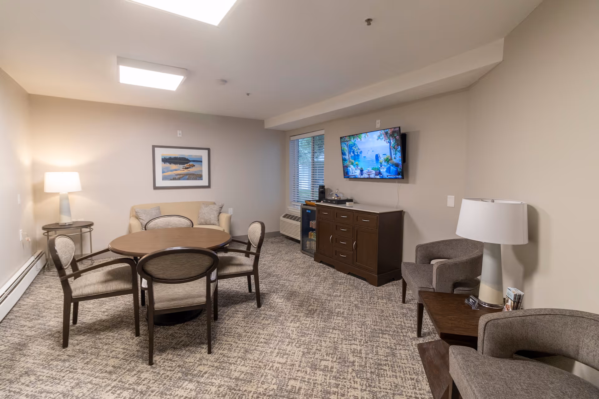 A cozy living room area with a round wooden table surrounded by four chairs in the center. There is a beige loveseat against the wall with two pillows and a framed landscape picture above it. On the right side, a flat-screen TV is mounted on the wall above a dark wooden cabinet with drawers and a mini fridge. Two gray armchairs flank a small wooden side table with a lamp and some brochures. The room has neutral-colored walls and carpeted flooring, with a window letting in natural light.