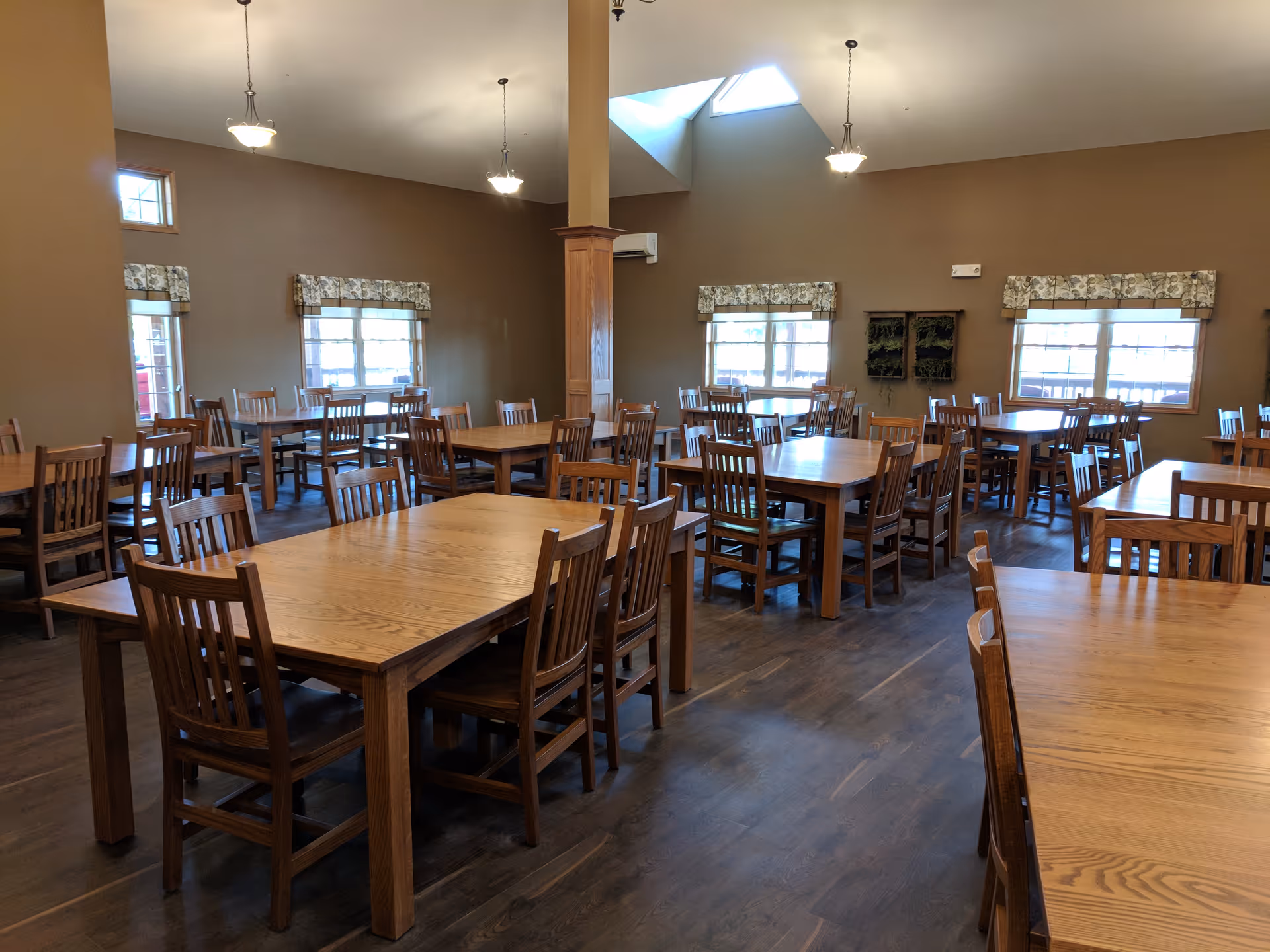 A spacious dining room with multiple wooden tables and chairs arranged neatly. The room has brown walls, several windows with floral valances, and hanging light fixtures. The floor is wooden, and there is a skylight providing natural light.