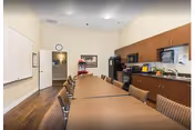 A conference or meeting room with a long wooden table surrounded by chairs. The room has a kitchenette area with cabinets, a microwave, a coffee maker, and a sink. There is a whiteboard on one wall and a clock above a door at the far end of the room. The floor is wooden, and the walls are light-colored.