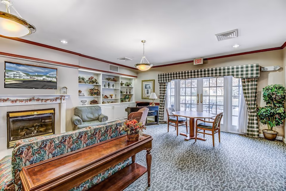 A cozy living room area in a senior living facility with floral patterned sofa, armchairs, a wooden coffee table with a flower arrangement, a fireplace with a TV mounted above it, built-in shelves with decorative items, a round wooden table with chairs near large glass doors leading to an outdoor patio, and green checkered curtains.