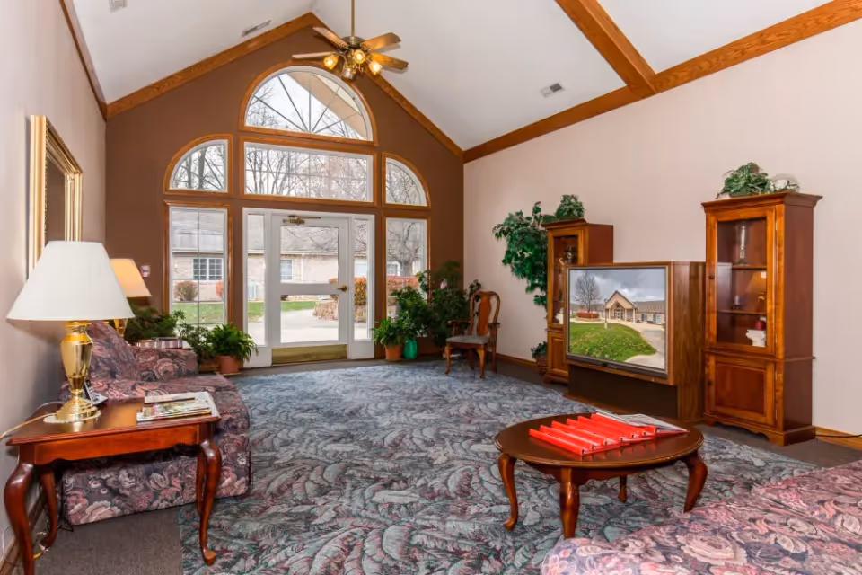 A cozy living room with floral patterned sofas, a wooden coffee table with red binders, a TV displaying an image of a building, wooden cabinets with plants and decorative items, large windows with an arched design letting in natural light, and a ceiling fan with lights.