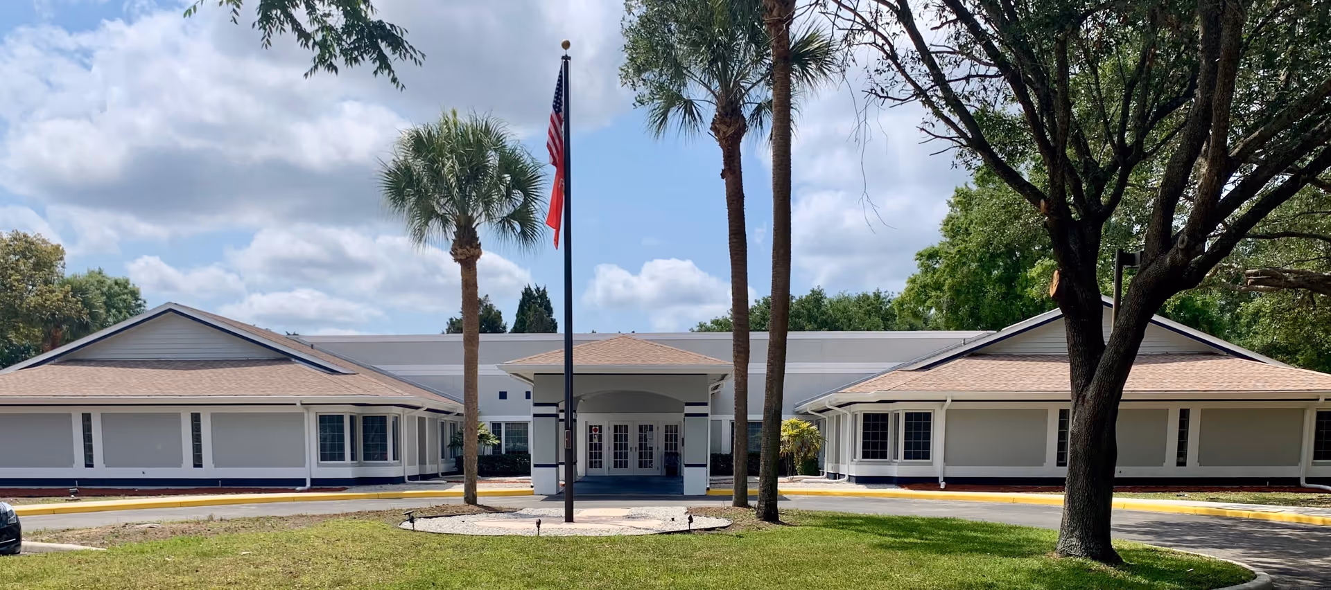 Front exterior view of a single-story assisted living facility building with a circular driveway, palm trees, an American flag, and a partly cloudy sky.
