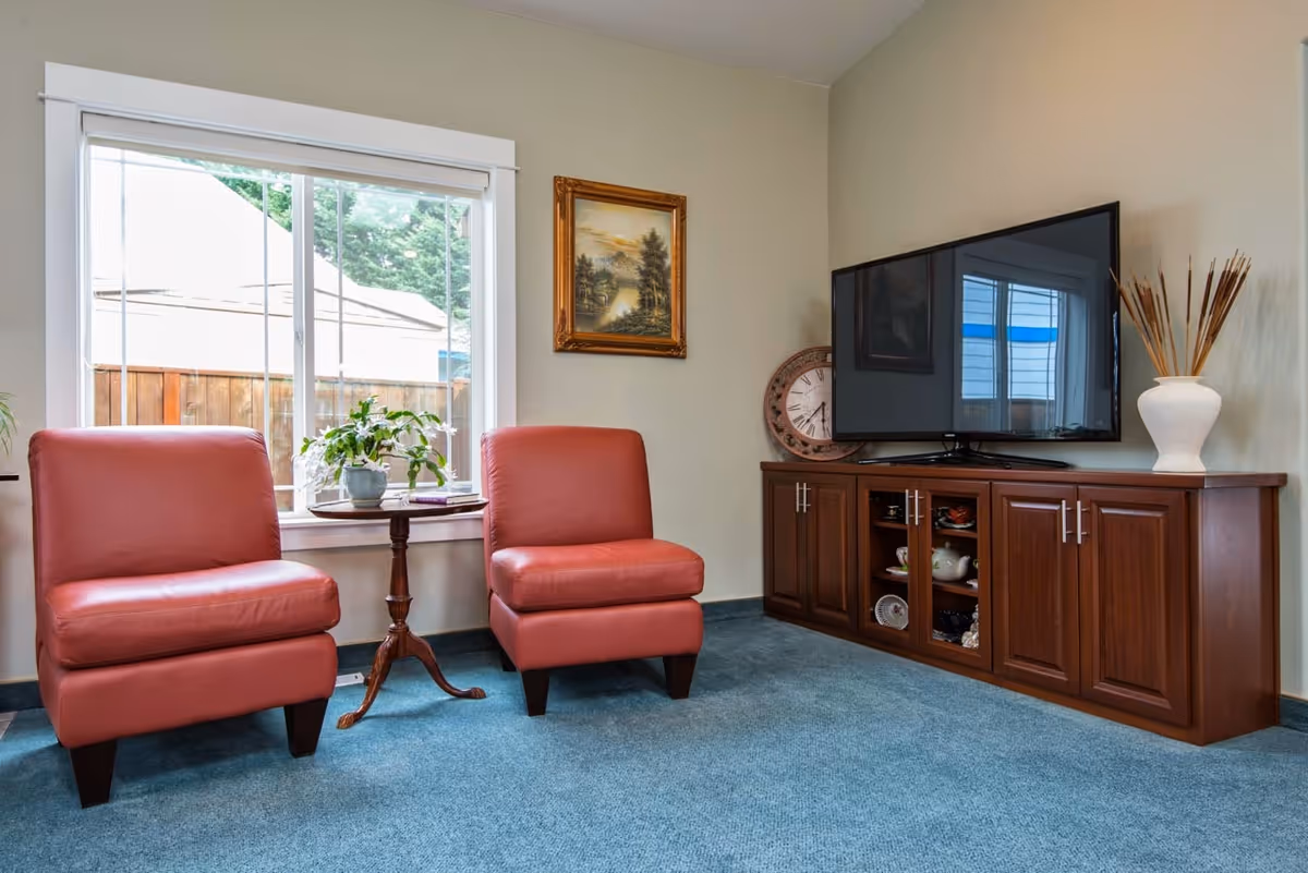 A cozy living room area with two red leather chairs separated by a small wooden side table holding a potted plant. There is a large window behind the chairs letting in natural light. On the right side, a wooden cabinet with glass doors holds decorative items and a large flat-screen TV sits on top. A framed landscape painting hangs on the wall above the cabinet, and a large decorative clock and white vase with dried plants are also on the cabinet.