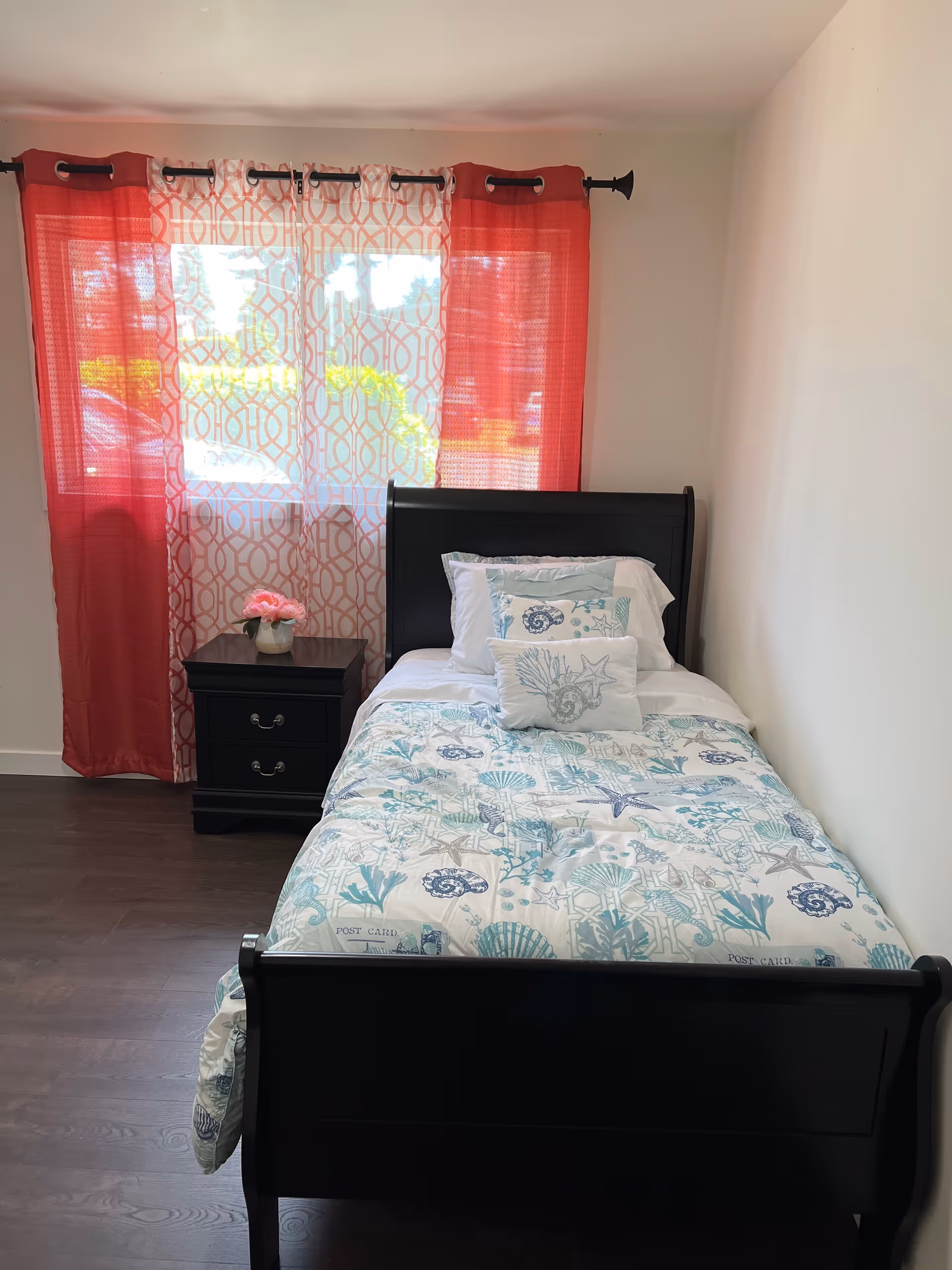 A small bedroom with a single black wooden bed against the right wall. The bed is made with white and blue seashell-themed bedding and pillows. To the left of the bed is a black nightstand with two drawers and a small vase of pink flowers on top. Behind the nightstand is a window with red and white patterned curtains allowing natural light into the room. The floor is dark wood and the walls are painted white.