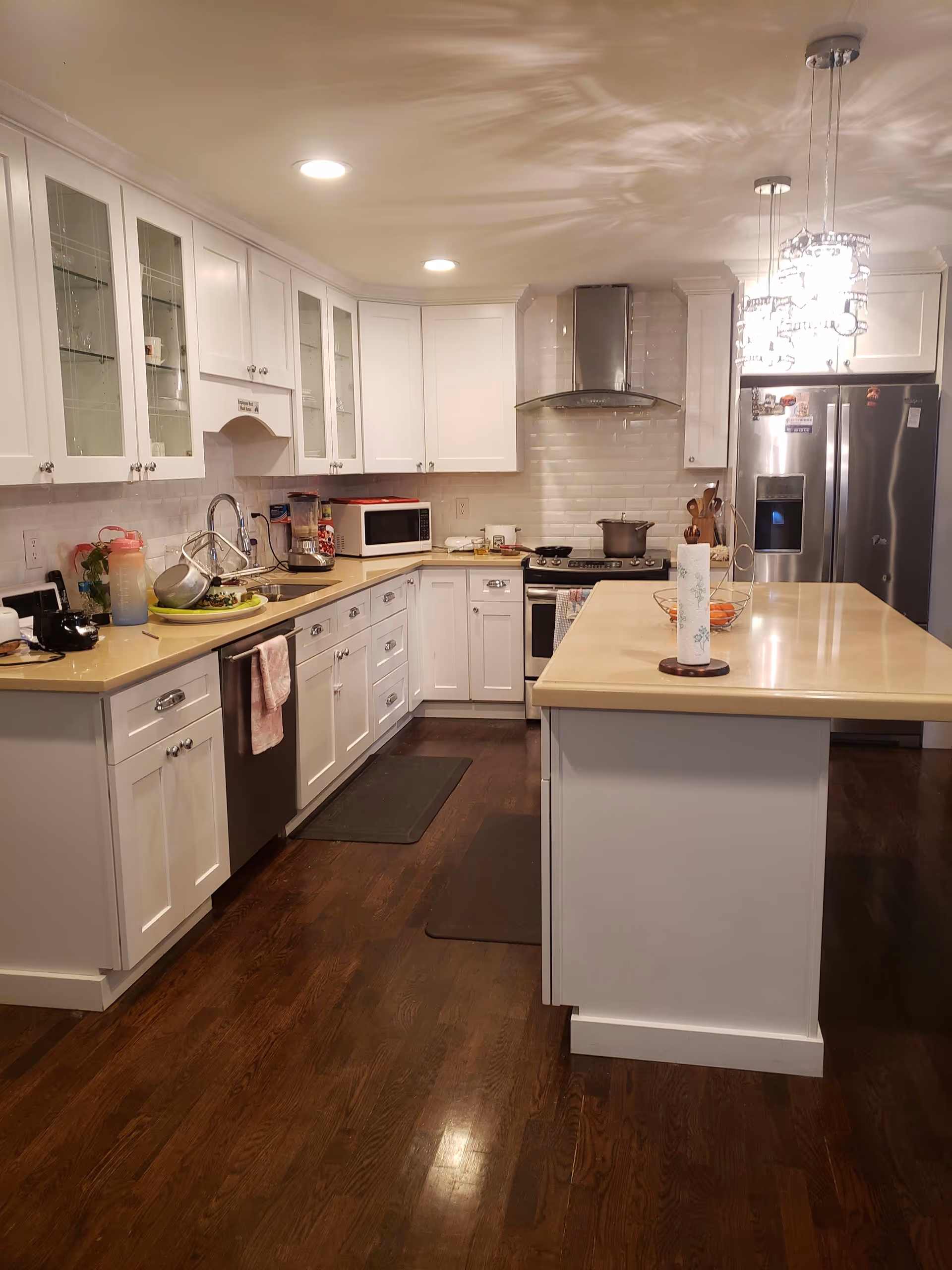 A modern kitchen with white cabinets and beige countertops. The kitchen features a central island with a paper towel holder and a fruit basket. Stainless steel appliances include a refrigerator, stove with a hood, dishwasher, and microwave. The floor is dark wood, and there are two pendant lights hanging above the island. Various kitchen items and utensils are placed on the counters.