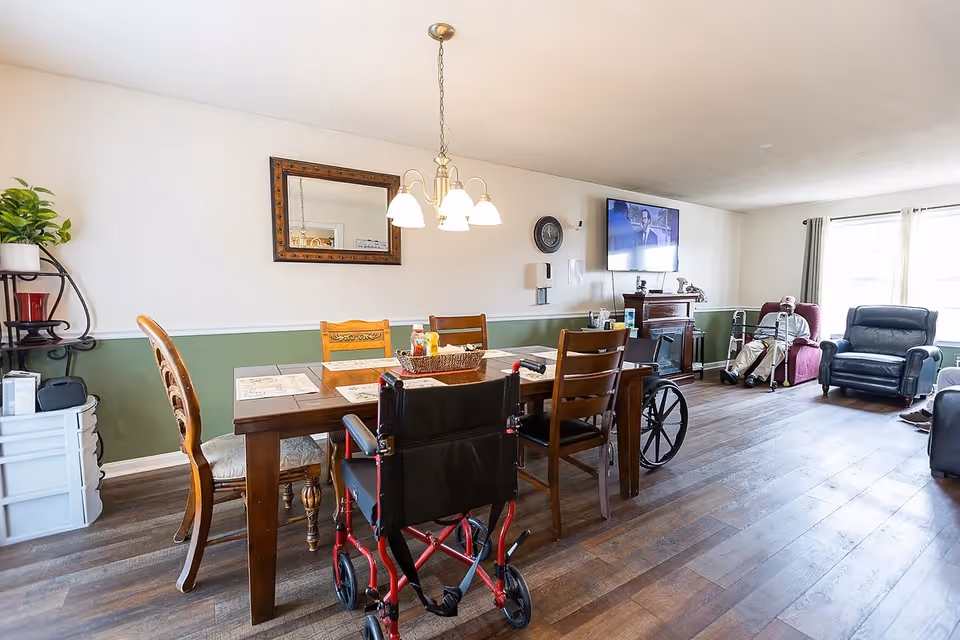 A bright and spacious common living area in a senior living facility featuring a wooden dining table with chairs and a wheelchair, a large wall-mounted TV above a fireplace, comfortable armchairs, and a large window letting in natural light. The walls are painted white with a green lower half, and there is a decorative mirror and a plant on a shelf.