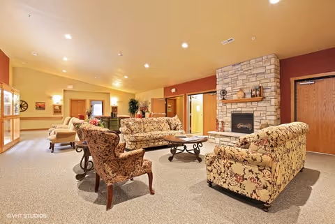 Spacious furnished lounge with floral sofas, armchairs, a coffee table and a stone fireplace in a senior living common area.