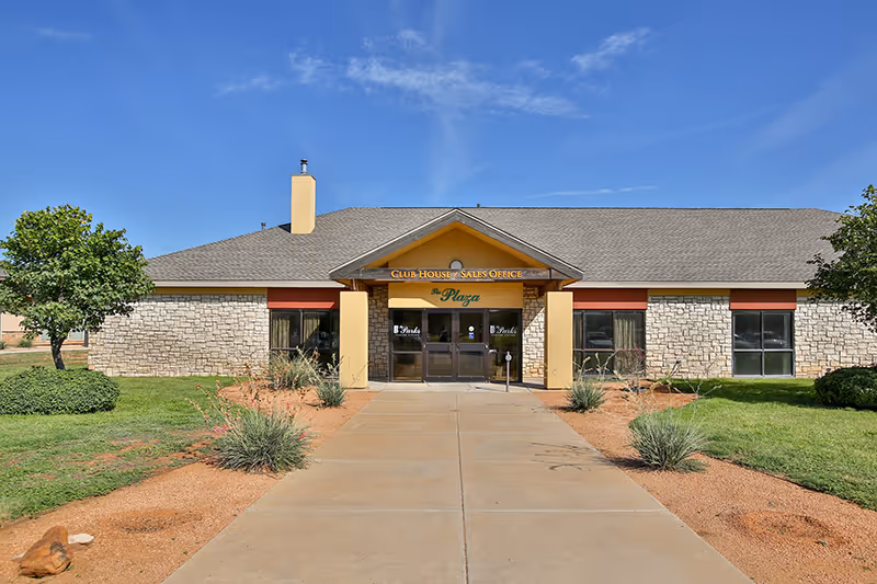 Exterior front view of a single-story building with stone and beige walls, a gray shingled roof, and a sign above the entrance that reads 'Club House Sales Office The Plaza'. The building is surrounded by green grass, small bushes, and a clear blue sky overhead.