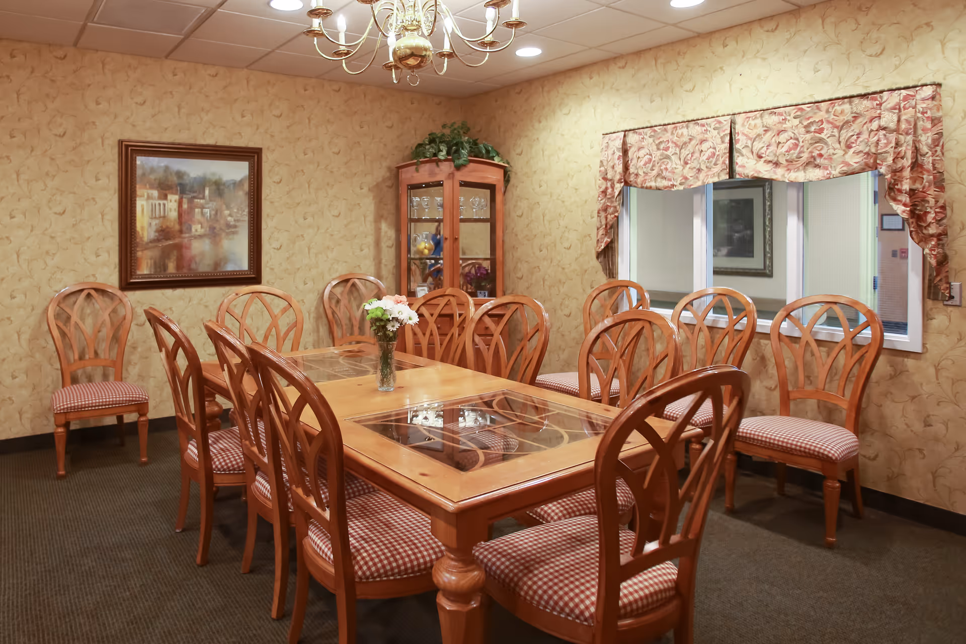 A dining room with a long wooden table surrounded by ten wooden chairs with red and white checkered cushions. A vase with flowers is placed in the center of the table. The walls are decorated with patterned wallpaper and a framed painting. A glass-fronted cabinet with glassware and decorative items is in the corner. A window with floral valance curtains is on one wall, and a chandelier hangs from the ceiling.
