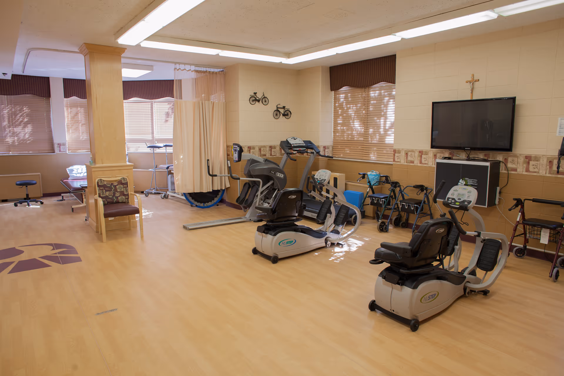 An indoor senior fitness and therapy room with recumbent exercise bikes, walkers, seating, a TV, and a privacy curtain.