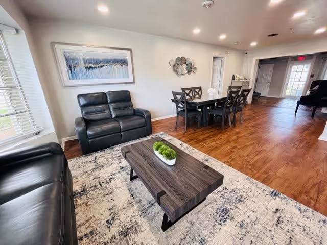 A bright living room with wooden flooring featuring a black leather loveseat and sofa, a wooden coffee table with a small white planter containing green plants, a patterned area rug, and a dining table with six chairs. The walls are light-colored with a framed abstract painting and decorative wall art. Large windows allow natural light to fill the room.