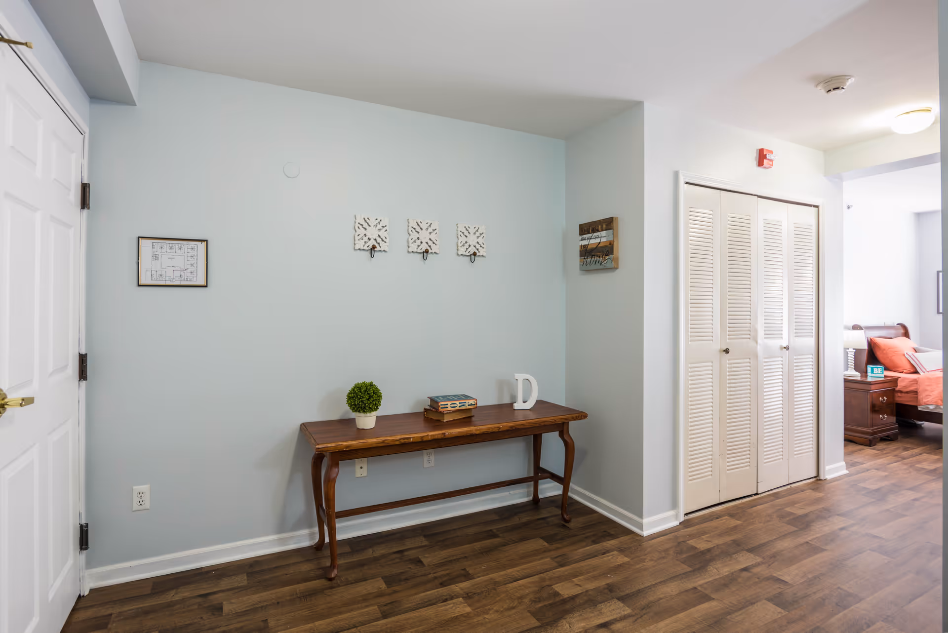 An interior hallway area with light blue walls and wood flooring. A wooden table against the wall holds a small potted plant, a book, and a decorative letter D. Above the table are three decorative wall hooks and two framed wall hangings. To the right, there is a white louvered closet door and a glimpse into a bedroom with a bed, nightstand, and orange bedding.