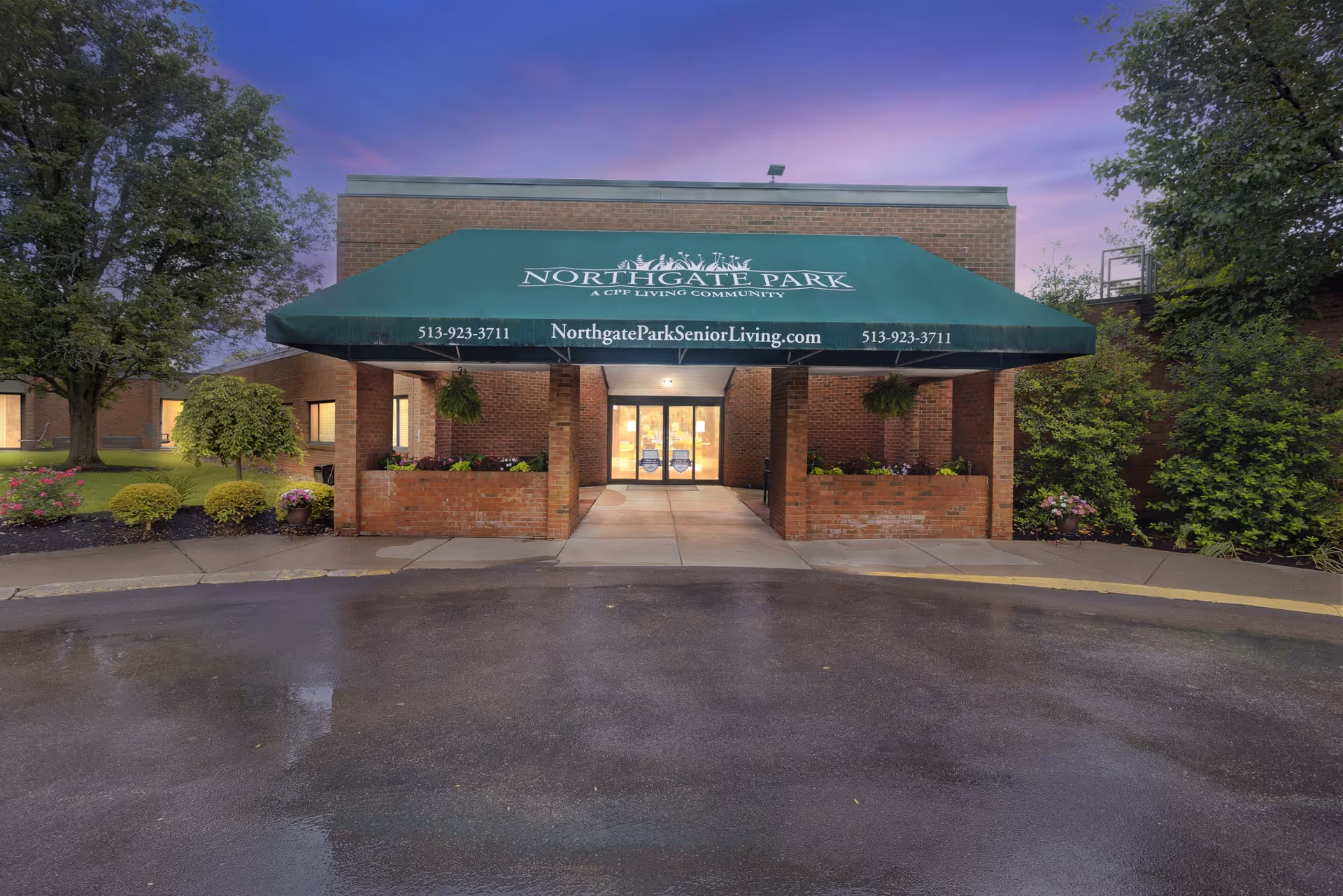 Entrance of Northgate Park senior living community with a green awning displaying the facility name, phone number, and website. The building is made of brick and is surrounded by trees and shrubs. The sky is purple and the ground appears wet.
