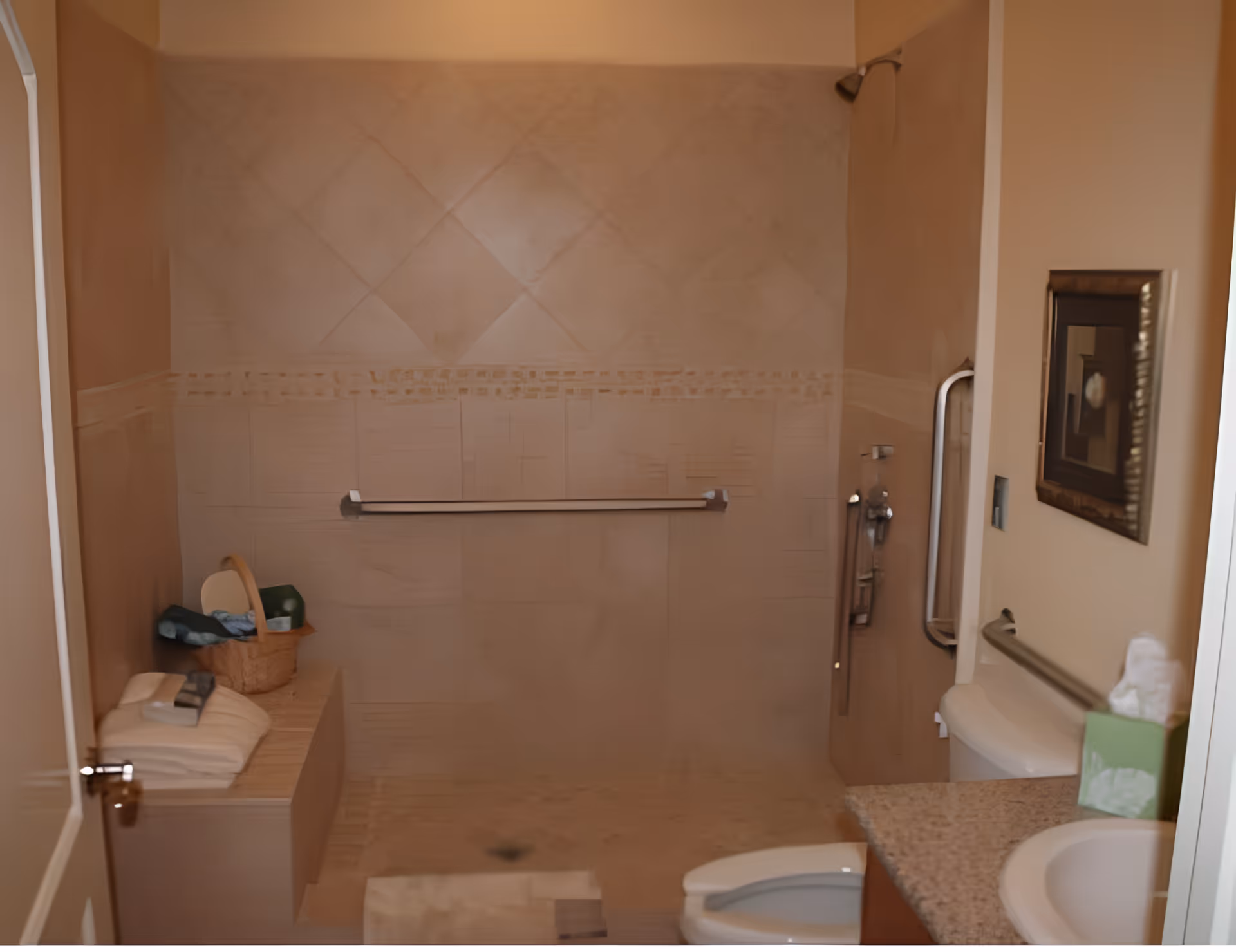 A bathroom with a walk-in shower featuring a grab bar, a bench with folded towels and a basket, a toilet, a sink with a granite countertop, a tissue box, and a framed picture on the wall.