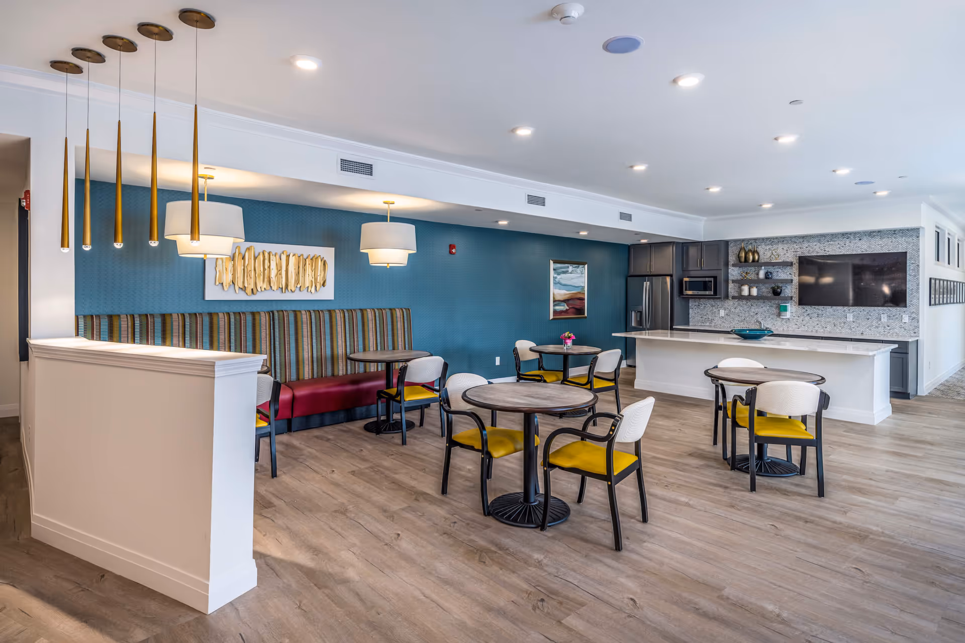 A modern dining area with round tables and chairs featuring yellow cushions. There is a striped bench along a teal accent wall with two pendant lights hanging above. The room also includes a kitchen area with a refrigerator, microwave, and a large countertop. The floor is wood, and the ceiling has recessed lighting.